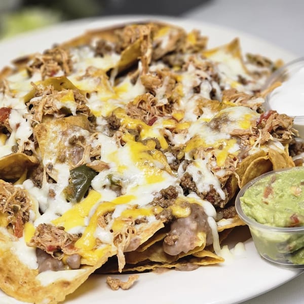A plate of loaded nachos with melted cheese, ground beef, shredded chicken, and a side of guacamole.
