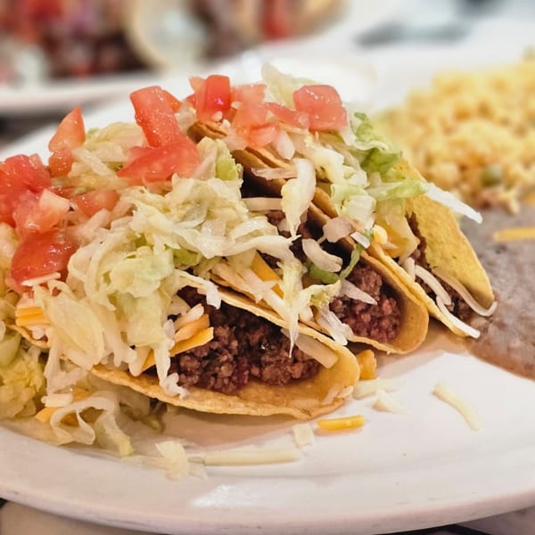 Three hard-shell tacos filled with ground beef, shredded lettuce, cheese, and diced tomatoes, with rice and beans.