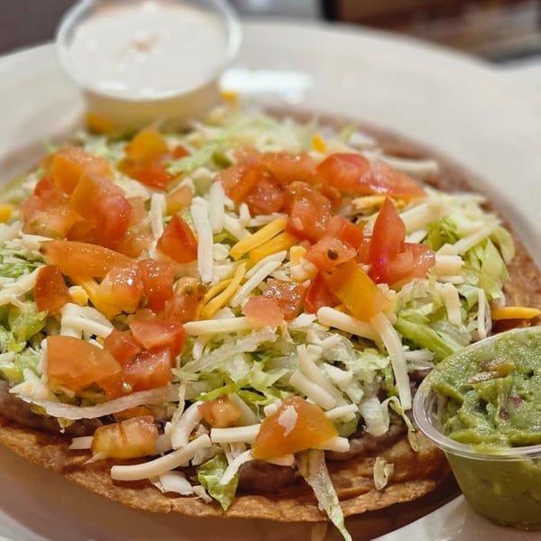 Crispy tostada topped with shredded lettuce, diced tomatoes, cheese, and a side of guacamole and sour cream.