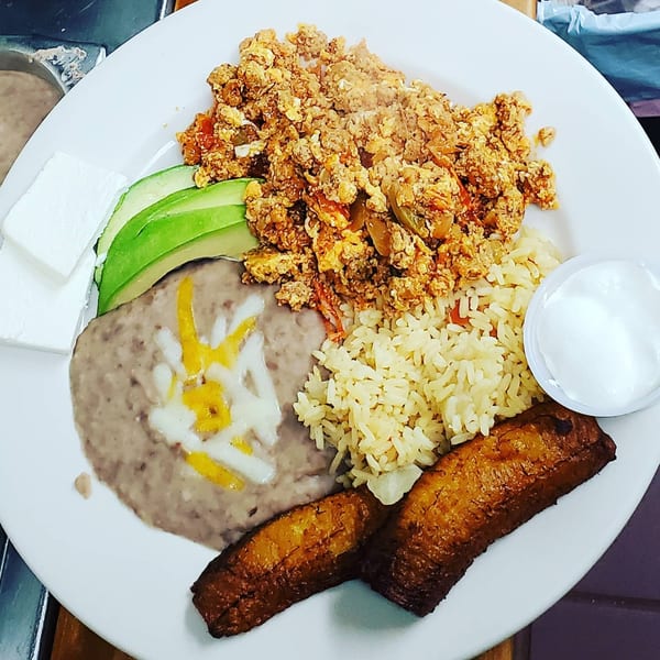 A Mexican breakfast plate with scrambled eggs, refried beans, rice, avocado, cheese, plantains, and sour cream.